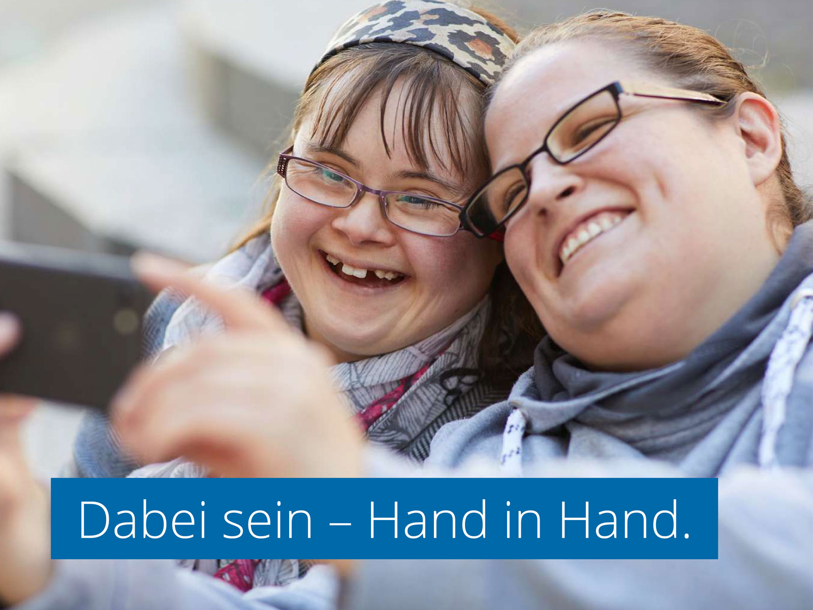 Zwei junge Frauen machen lachend ein Selfie. Eingelassener Text: "Dabei sein - Hand in Hand."