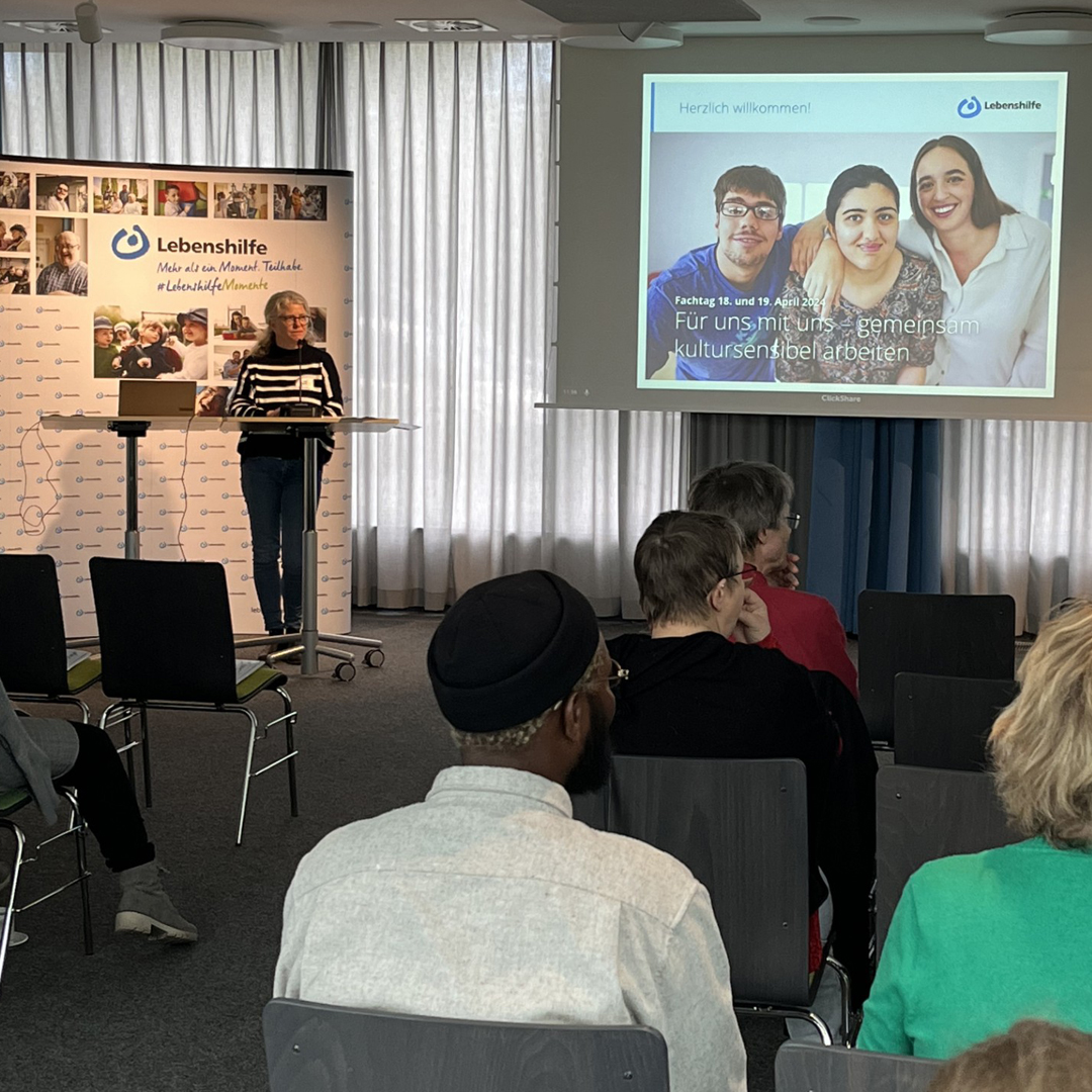 Fachtag Für uns mit uns am 18 udn 19. April 2024. Begrüßung durch Antje Welke. Im Vordergrund sitzen Personen im Publikum. Sie schauen auf eine Leinwand und eine weibliche Person, die an einem Rednerpult steht. d