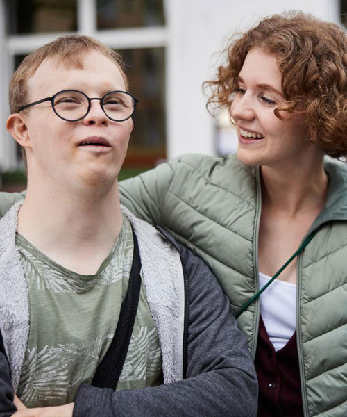Ein junger Mann mit Behinderung neben einer jungen Frau, die ihn freundlich anlächelt.