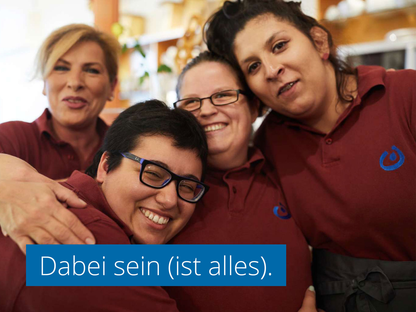 Vier Frauen in gleichen T-Shirts (offenbar das Team eines Cafés) lachen und umarmen sich. Eine Schrift ist in das Foto eingelassen: "Dabei sein (ist alles)."