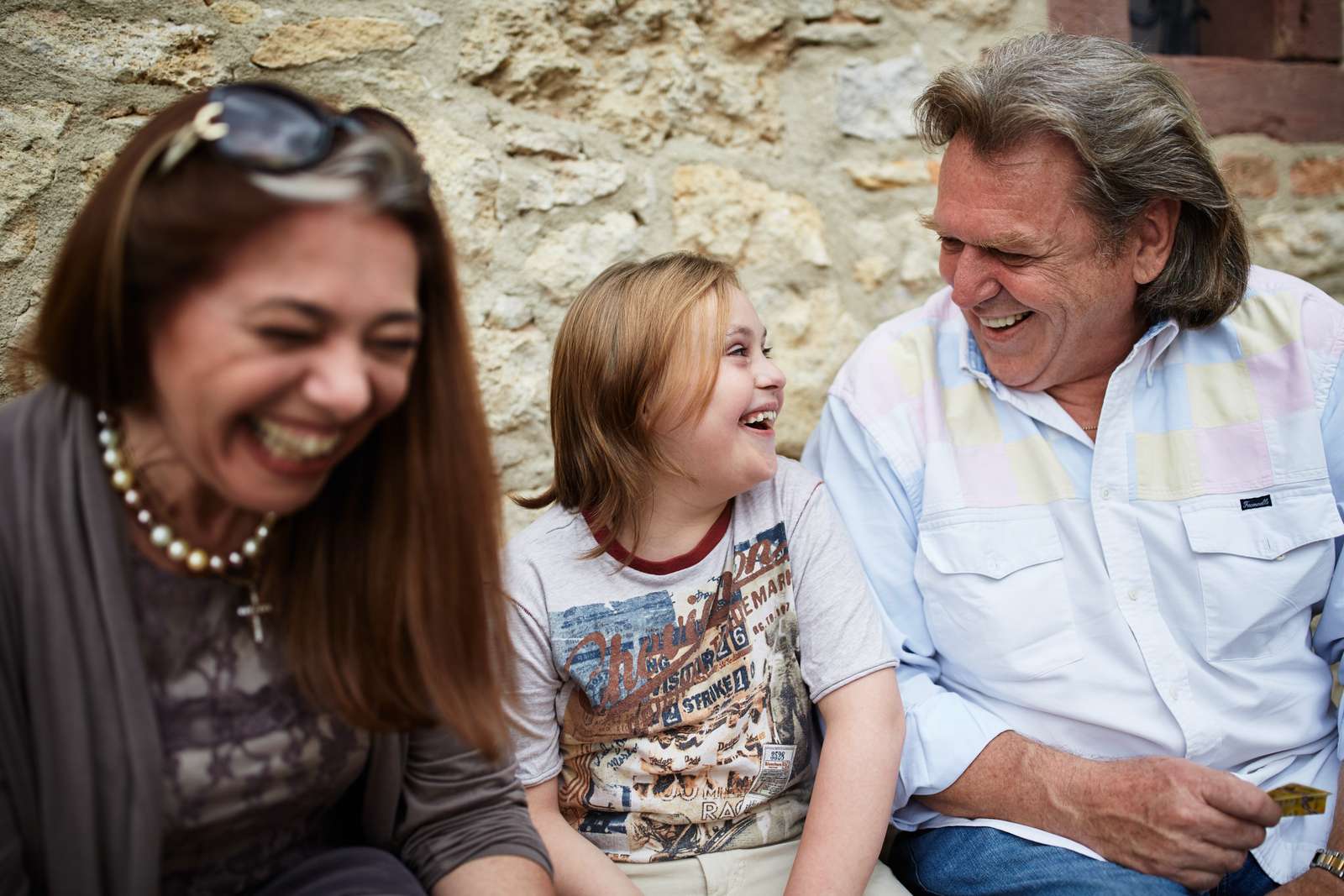 Eine Frau, ein Mann und ein Kind sitzen zusammen vor einer Mauer und lachen zusammen, lauthals.