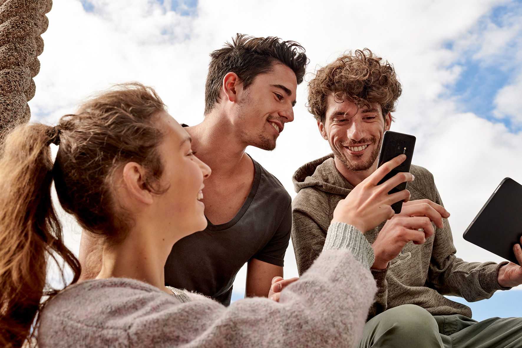 Drei junge Menschen (eine Frau, zwei Männer) schauen gemeinsam auf ein Handy und ein Tablet und lachen fröhlich dabei.