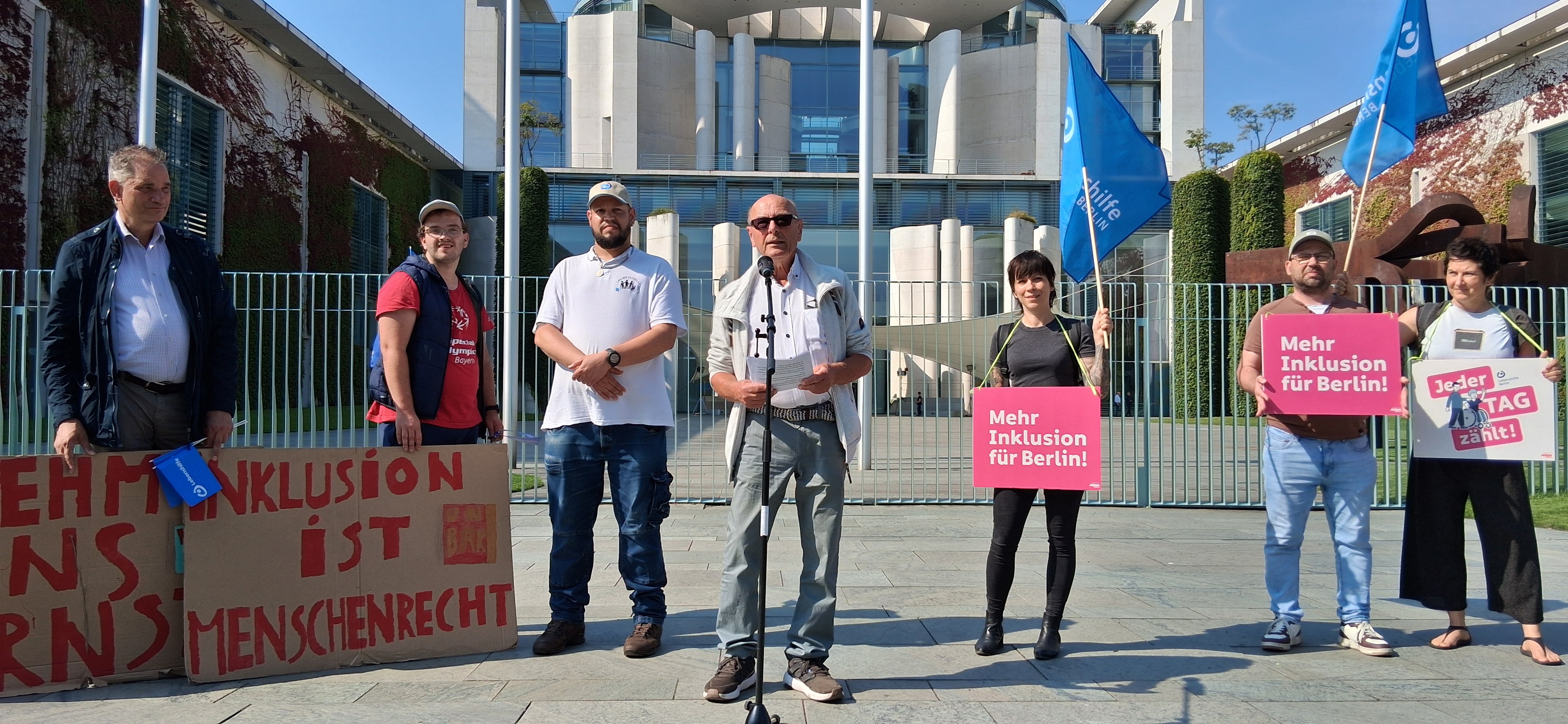 Rolf Flathmann, Stellvertretender Bundesvorsitzender, spricht auf der Demo für die Petition "Teilhabe ist kein Luxus" vorm Bundeskanzleramt. Die weiteren Personen auf dem Bild, von links:Christian Keitmann, Marcus Dengler, Philipp Grossmann (alle drei Selbstvertreter der Lebenshilfe Altmühlfranken), Katja Macheleidt, Büro für Selbstvertretung der Lebenshilfe Berlin, Sascha Ubrig, hauptamtlicher Selbstvertreter der Lebenshilfe Berlin, und Anja Hahlweg, Büro für Selbstvertretung der Lebenshilfe Berlin.
