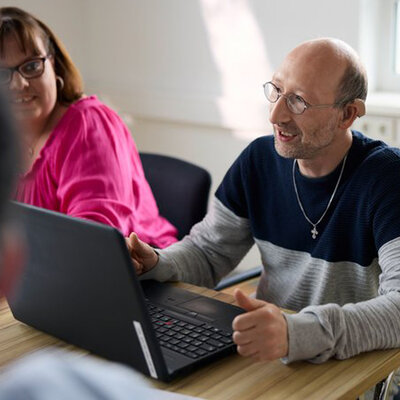 Über den Begriff "Behinderung" Auf dem Bild ist ein Mann mit sogenannter geistiger Behinderung zu sehen. Er sitzt vor einem Laptop und spricht gerade.