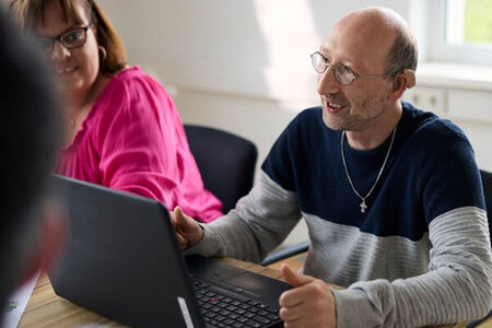 Auf dem Bild ist ein Mann mit sogenannter geistiger Behinderung zu sehen. Er sitzt vor einem Laptop und spricht gerade.