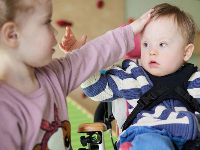 Ein kleines Mädchen streichelt einen Jungen, der in einem Rollstuhl sitzt. Der Junge hat eine sogenannte geistige Behinderung.