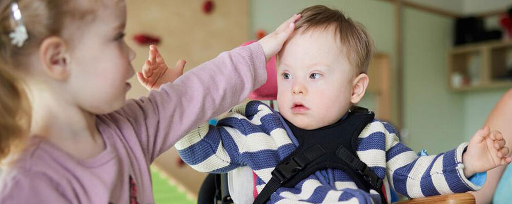 Ein kleines Mädchen streichelt einen Jungen, der in einem Rollstuhl sitzt. Der Junge hat eine sogenannte geistige Behinderung.