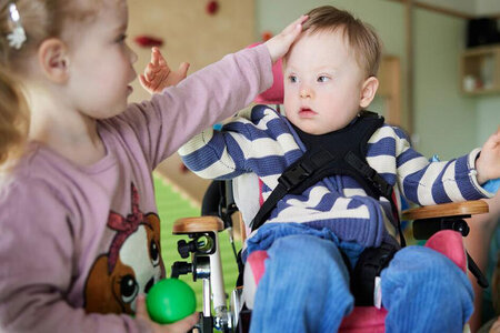 Ein kleines Mädchen streichelt einen Jungen, der in einem Rollstuhl sitzt. Der Junge hat eine sogenannte geistige Behinderung.
