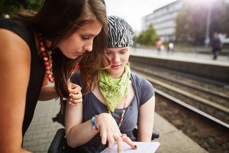 Zwei junge Frauen sind zusammen am Bahnsteig, eine davon im Rollstuhl. Sie schauen gemeinsam in einen Fahrplan.