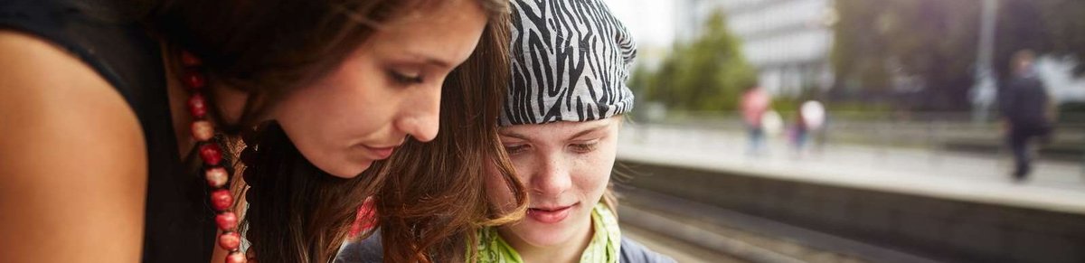 Zwei junge Frauen sind zusammen am Bahnsteig, eine davon im Rollstuhl. Sie schauen gemeinsam in einen Fahrplan.