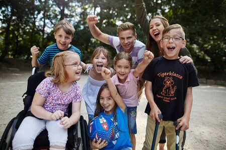 Gruppe von Kindern auf dem Schulhof. Sie haben teils eine Behinderung. Alle lachen in die Kamera oder jubeln mit erhobenen Armen.
