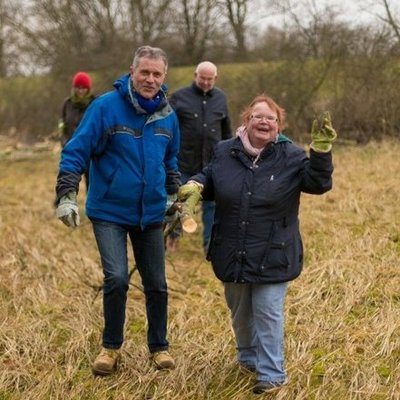 Ein Mann und eine Frau tragen zusammen Geäst. Sie tragen Arbeitshandschuhe und lachen strahlend. Im Hintergrund weitere Helfer.