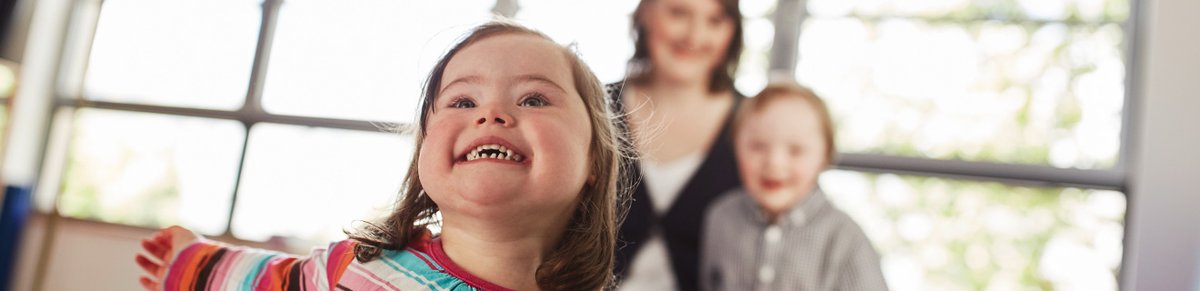 Zwei Kinder mit Down-Syndrom lächeln. Im Hintergrund steht eine Frau.