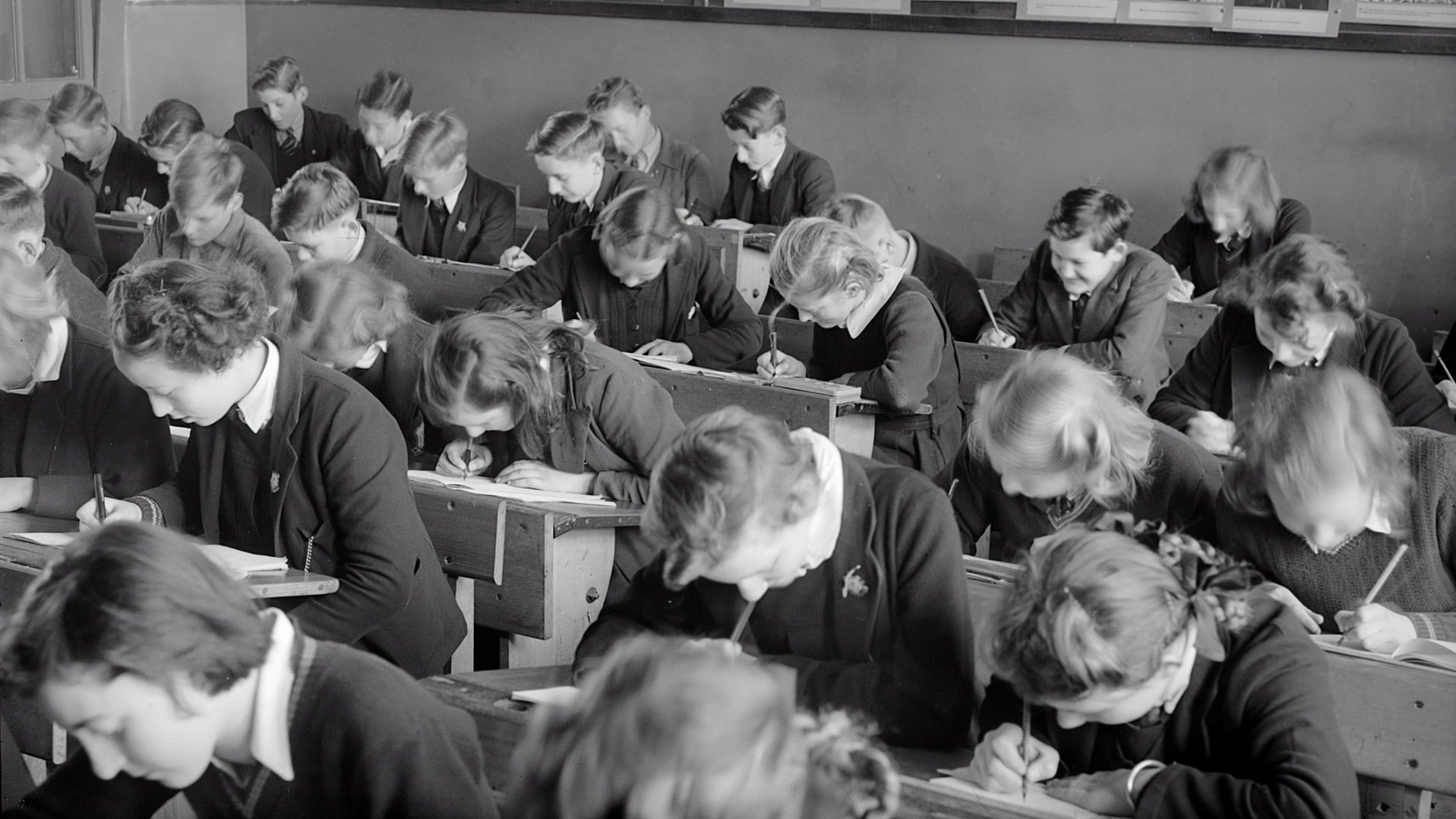 Klassenzimmer mit Schülerinnen und Schülern in Bänken, die über ihre Bücher und Hefte gebeugt sind. Das Foto ist in schwarz/weiß.