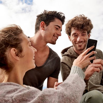 Drei junge Menschen (eine Frau, zwei Männer) schauen gemeinsam auf ein Handy und ein Tablet und lachen fröhlich dabei.