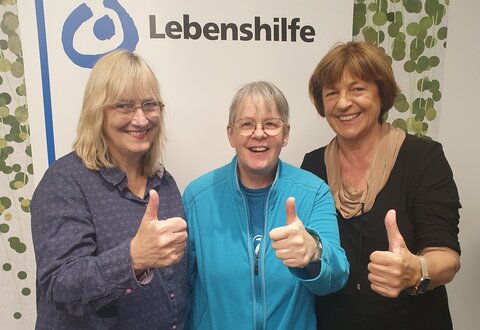 Von rechts: Ulla Schmidt, Bundesvorsitzende der Lebenshilfe, Ramona Günther, Mitglied im Bundesvorstand und im Rat behinderter Menschen der Lebenshilfe, sowie Bundesgeschäftsführerin Jeanne Nicklas-Faust, freuen sich über die Spende der NÜRNBERGER Versicherung. Alle drei zeigen einen Daumen hoch.