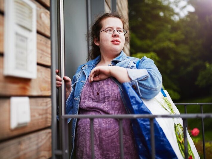 Behinderung oder Beeinträchtigung? Eine junge Frau steht vor einer Wohnungstür. Sie hat eine sogenannte geistige Beeinträchtigung.
