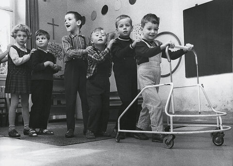 Mehrere fröhliche Kinder mit Behinderung stehen hintereinander in einem Lebenshilfe-Kindergarten in Heidelberg, 1968.