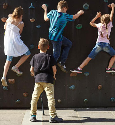 Was ist Exklusion? Ein Junge mit Beeinträchtigung steht allein vor einer Kletterwand, während andere Kinder hochklettern.