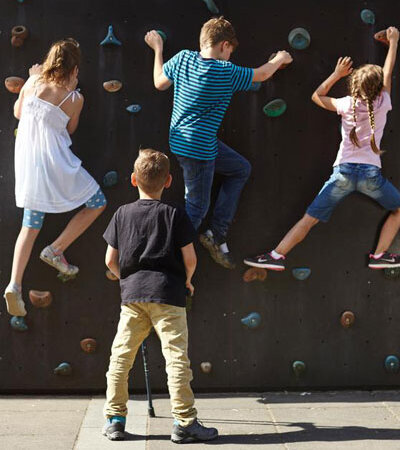 Ein Junge mit Beeinträchtigung steht allein vor einer Kletterwand, während andere Kinder hochklettern.