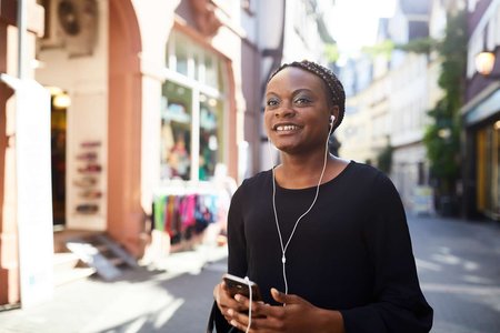 Eine lächelnde junge Frau mit Handy und In-Ear-Kopfhörern läuft eine schmale Straße mit Geschäften und Wohnhäusern entlang.