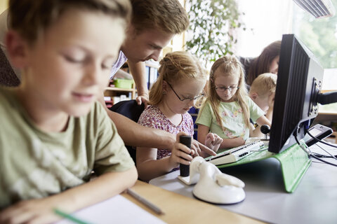 Das Bild zeigt mehrere Schüler*innen im Unterricht in einer Tischreihe. Mitten auf dem Tisch steht ein Computer-Bildschirm, an dem eine Schülerin mit Behinderung vom Lehrer unterstützt wird.