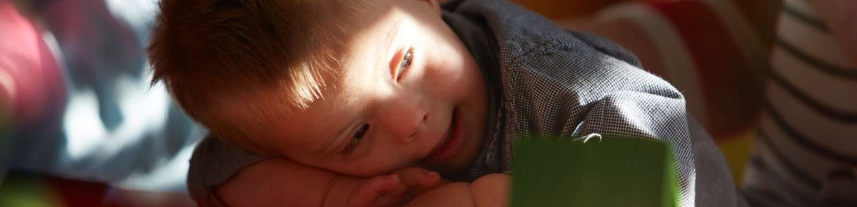 Ein kleiner Junge mit Down-Syndrom spielt mit Bauklötzchen. Es fällt ein warmer Lichtstrahl aus einem Fester auf ihn.  