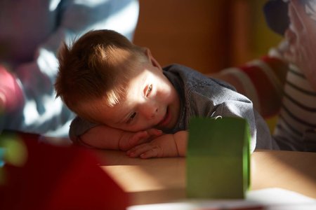 Ein kleiner Junge mit Down-Syndrom spielt mit Bauklötzchen. Es fällt ein warmer Lichtstrahl aus einem Fester auf ihn.  