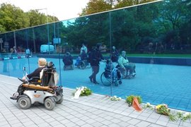 Das Bild zeigt den Gedenk- und Informationsort zu den "Euthanasie"-Verbrechen der Nationalsozialisten in der Berliner Tiergartenstraße. Zu sehen sind eine durchsichtige blaue Wand, Rollstuhlfahrer und Fußgänger sowie Blumen und Kränze, die auf dem Boden liegen.