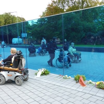 Das Bild zeigt den Gedenk- und Informationsort zu den "Euthanasie"-Verbrechen der Nationalsozialisten in der Berliner Tiergartenstraße. Zu sehen sind eine durchsichtige blaue Wand, Rollstuhlfahrer und Fußgänger sowie Blumen und Kränze, die auf dem Boden liegen.