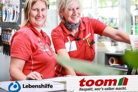 Auf dem Foto sind die Logos von toom und Lebenshilfe zu sehen und zwei Baumarkt-Mitarbeiterinnen in roten Hemden.
