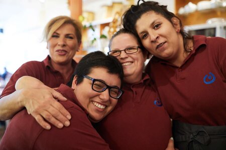 Vier lachende Frauen mit und ohne Behinderung. Sie tragen alle die Mitarbeiterkleidung eines Cafés: weinrote T-Shirts mit Lebenshilfe-Logo.