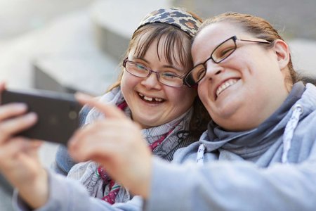 Zwei junge Frauen stecken die Köpfe zusammen, machen ein Selfie und lachen dabei über das ganze Gesicht.