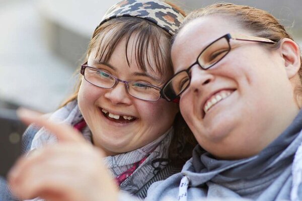 Zwei junge Frauen stecken die Köpfe zusammen, machen ein Selfie und lachen dabei über das ganze Gesicht.